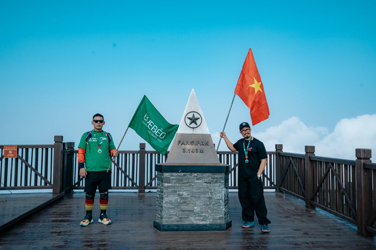 Hai người trưởng thành đứng cạnh cột mốc Fansipan, cầm cờ Bye Béo và cờ Việt Nam.